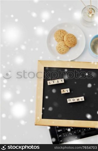 quarantine, epidemic and safety concept - close up of chalkboard with stay at home words on wooden toy blocks, coffee cup, cookies and book on white background in winter over snow. chalkboard with stay at home words on toy blocks