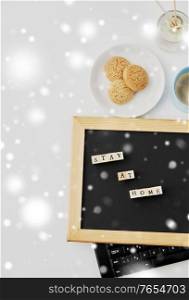quarantine, epidemic and safety concept - close up of chalkboard with stay at home words on wooden toy blocks, coffee cup, cookies and book on white background in winter over snow. chalkboard with stay at home words on toy blocks