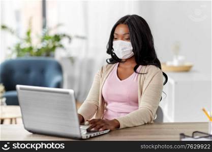 quarantine, remote job and pandemic concept - african american woman wearing face protective medical mask for protection from virus disease with laptop computer working at home office. woman in mask with laptop working at home office