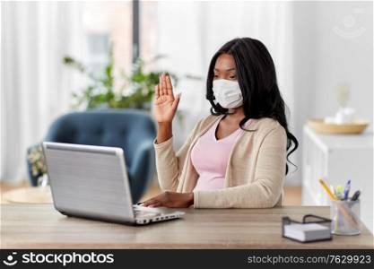 quarantine, remote job and pandemic concept - african american woman wearing face protective medical mask for protection from virus disease with laptop computer having video call at home office. woman in mask with laptop has video call at home