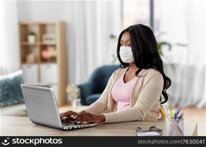 quarantine, remote job and pandemic concept - african american woman wearing face protective medical mask for protection from virus disease with laptop computer working at home office. woman in mask with laptop working at home office