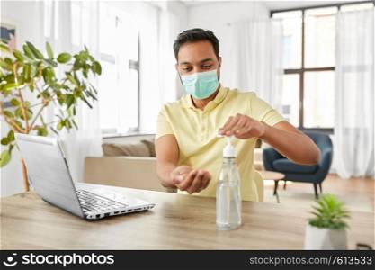 quarantine, remote job and pandemic concept - happy indian man wearing face protective medical mask for protection from virus disease with laptop computer using hand sanitizer at home office. man in mask using hand sanitizer at home office