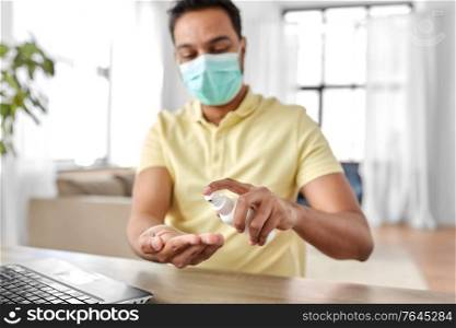 quarantine, remote job and pandemic concept - happy indian man wearing face protective medical mask for protection from virus disease using hand sanitizer with laptop computer working at home office. man in mask using hand sanitizer at home office