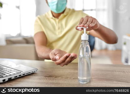 quarantine, remote job and pandemic concept - happy indian man wearing face protective medical mask for protection from virus disease using hand sanitizer with laptop computer working at home office. man in mask using hand sanitizer at home office