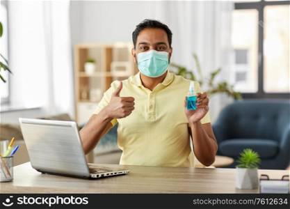 quarantine, remote job and pandemic concept - indian man wearing face protective medical mask for protection from virus disease with hand sanitizer and laptop computer showing thumbs up at home office. man in mask with hand sanitizer at home office