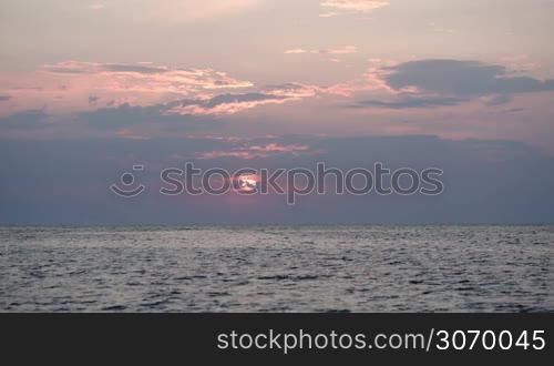 Quiet sea at sunset, sun almost hidden with clouds, bird flying over the water