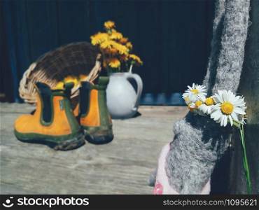 rabbit toy with flowers wreath on head, yellow flowers and rubber gardening ankle boots on wooden veranda background. Still life in rustic style. Daylight, hard shadows. Countryside lifestyle fun concept