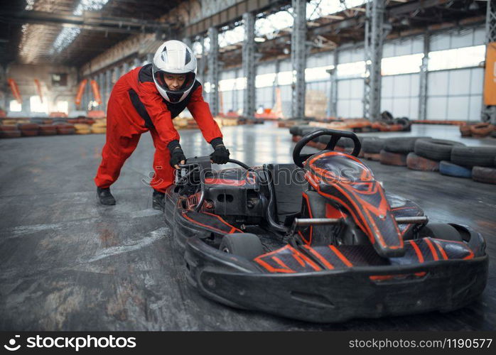 Racer in helmet pushing a go kart car, karting auto sport indoor. Speed race on close go-kart track with tire barrier. Fast vehicle competition, high adrenaline leisure