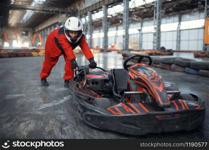 Racer in helmet pushing a go kart car, karting auto sport indoor. Speed race on close go-kart track with tire barrier. Fast vehicle competition, high adrenaline leisure