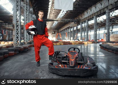 Racer with helmet poses near go kart car, karting auto sport indoor. Speed race on close go-kart track with tire barrier. Fast vehicle competition, high adrenaline leisure