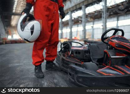 Racer with helmet poses near go kart car, karting auto sport indoor. Speed race on close go-kart track with tire barrier. Fast vehicle competition, high adrenaline leisure. Racer with helmet poses near go kart car, karting