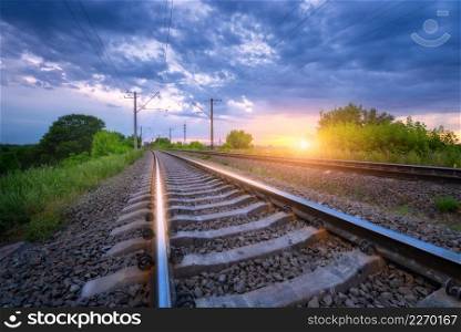 Railroad and beautiful blue sky at golden sunset in summer. Railway station and sky with clouds. Industrial landscape with railway platform, green trees and grass, dramatic cloudy sky in the evening