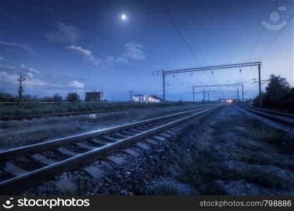 Railroad and blue sky with moon and clouds at night. Summer rural industrial landscape with railway station, trees, grass at twilight. Railway platform at dusk. Freight transportation. Heavy industry. Railroad and blue sky with moon and clouds at night