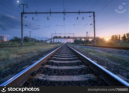 Railroad and blue sky with moon at sunset. Summer rural industrial landscape with railway station, sky with clouds and sunlight, green grass. Railway platform. Transportation. Heavy industry at dusk. Summer rural industrial landscape with railway station at sunset