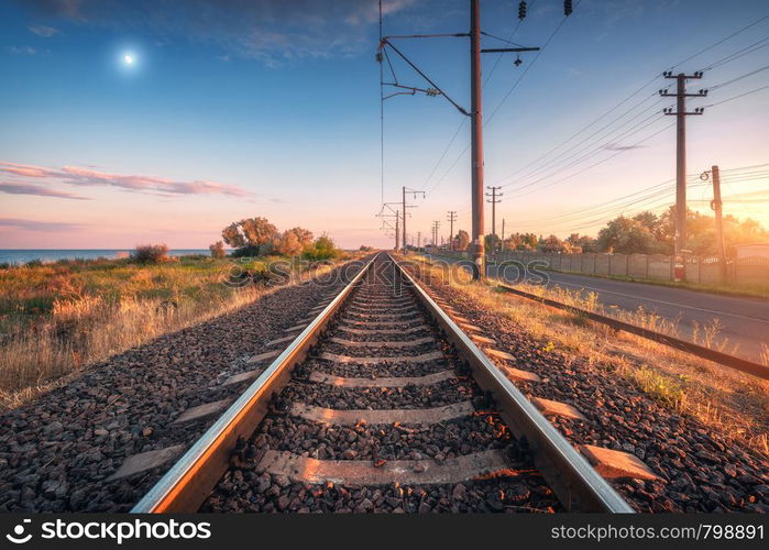 Railroad and blue sky with moon at sunset. Summer rural industrial landscape with railway station, sky with clouds and gold sunlight, green grass. Railway platform. Transportation. Heavy industry. Railroad and blue sky with moon at sunset in summer