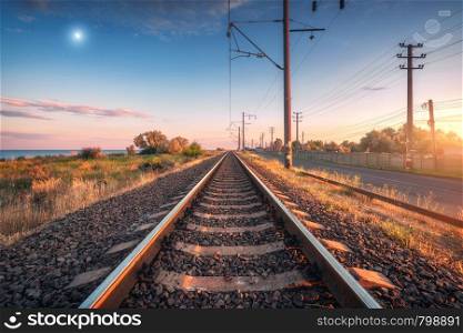 Railroad and blue sky with moon at sunset. Summer rural industrial landscape with railway station, sky with clouds and gold sunlight, green grass. Railway platform. Transportation. Heavy industry. Railroad and blue sky with moon at sunset in summer