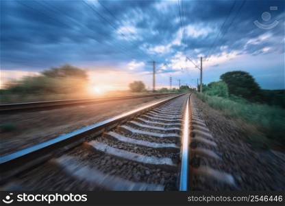 Railroad and dramatic cloudy blue sky at sunset with motion blur effect in summer. Railway station in motion. Industrial landscape with blurred railway platform, green trees and grass in the evening