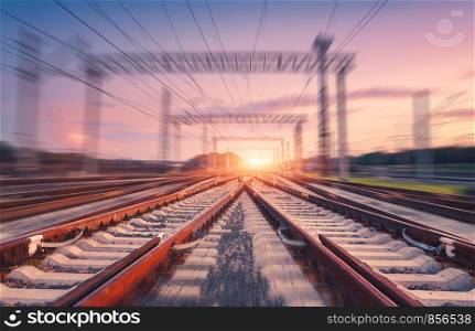 Railroad and pink sky with motion blur effect at sunset. Industrial landscape with railway station, light and blurred background at twilight. Railway platform in move. Transportation. Speed motion. Railroad and pink sky with motion blur effect at sunset