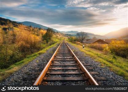 Railroad in mountains at sunset in autumn. Beautiful industrial landscape with railway station, orange trees, green grass, buildings, rocks and blue sky with clouds in fall. Rural railway platform