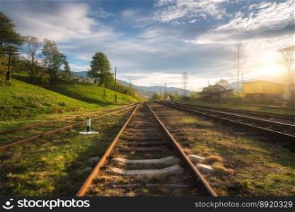 Railroad in mountains at sunset in summer. Beautiful industrial landscape with railway station, trees, green grass, blue sky, clouds in spring. Old rural railway platform in Ukraine. Transportation. Railroad in mountains at sunset in summer. Industrial landscape