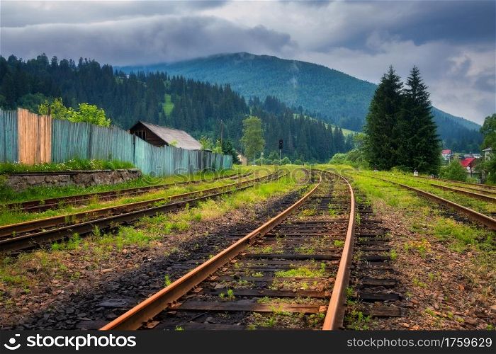 Railroad in mountains in overcast summer day. Industrial landscape with old railway station, trees, green grass, buildings, hills in fog, moody sky with clouds in fall. Railway platform in village
