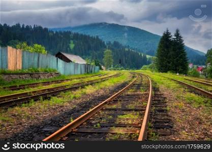 Railroad in mountains in overcast summer day. Industrial landscape with old railway station, trees, green grass, buildings, hills in fog, moody sky with clouds in fall. Railway platform in village