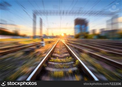 Railroad with motion blur effect at sunset. Industrial conceptual landscape with blurred railway station, buildings, green grass, blue sky and orange sunlight in the evening. Railway track. Background. Railroad with motion blur effect at sunset
