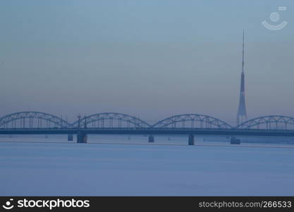 Railway bridge and television tower in winter. Railway bridge over the frozen river Daugava in Riga.TV tower and railway bridge on blue sky background in winter. Riga view in winter.