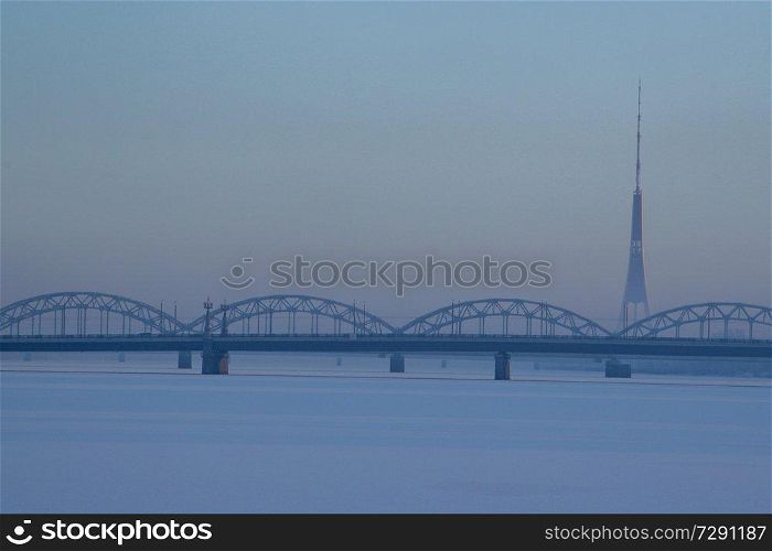 Railway bridge and television tower in winter. Railway bridge over the frozen river Daugava in Riga.TV tower and railway bridge on blue sky background in winter. Riga view in winter.