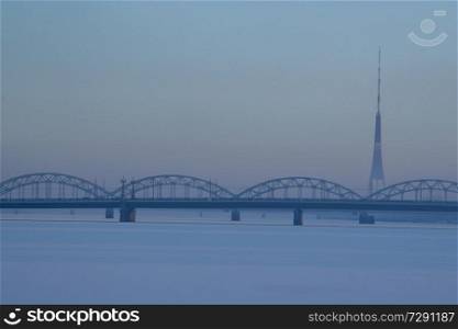 Railway bridge and television tower in winter. Railway bridge over the frozen river Daugava in Riga.TV tower and railway bridge on blue sky background in winter. Riga view in winter.