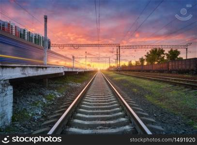 Railway station with blurred high speed train and freight train at colorful sunset. Railroad in summer. Industrial landscape with moving train, railway platform, sky with pink clouds. Transportation