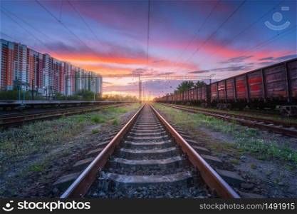 Railway station with freight trains and multicolored buildings at sunset. Railroad in summer. Heavy industry. Landscape with train, railway platform, sky with colorful clouds at dusk. Transportation
