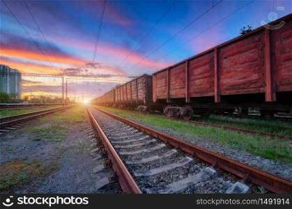 Railway station with freight trains at colorful sunset. Railroad in summer. Heavy industry. Industrial landscape with train, green grass, railway platform, blue sky with pink clouds. Transportation