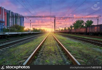 Railway station with freight trains at colorful sunset. Railroad in summer. Heavy industry. Industrial landscape with train, green grass, railway platform, purple sky with pink clouds. Transportation