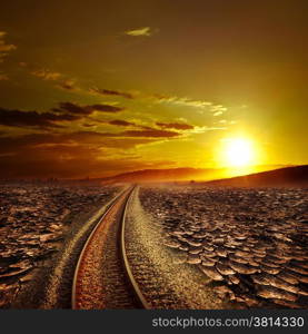 Railway track crossing drought cracked desert landscape under dramatic evening sunset sky. Global warming and travel concept