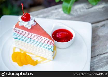 Rainbow crepe cake and a cherry with strawberry sauce on wooden table background.