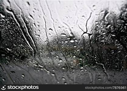 Raindrops on a glass window with blurry trees, road and gray car
