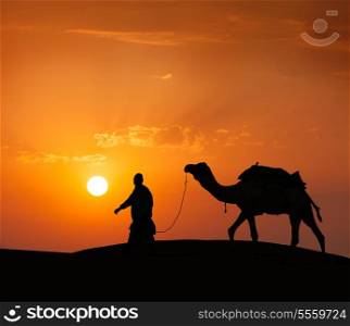 Rajasthan travel background - indian cameleer (camel driver) with camel silhouette in dunes of Thar desert on sunset. Jaisalmer, Rajasthan, India