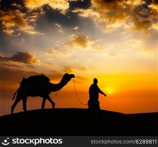 Rajasthan travel background - indian cameleer camel driver with camel silhouette in dunes of Thar desert on sunset. Jaisalmer, Rajasthan, India