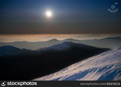 Range of winter mountains in white snow at night under dark blue sky with moon and stars