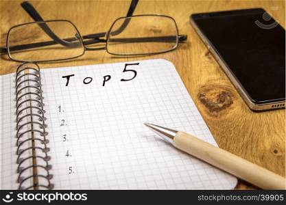 Ranking concept for a top 5 preferences list, written on a graph, spiral notebook sheet, placed on a wooden table, with eyeglasses and a phone in the background.