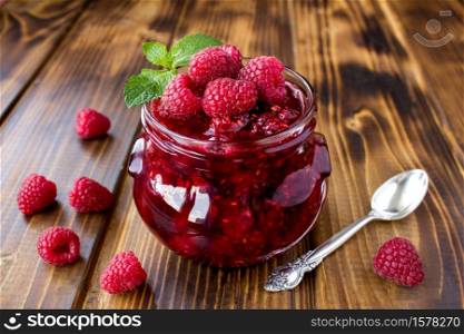 Raspberry jam in the glass jar on the brown wooden background. Closeup.