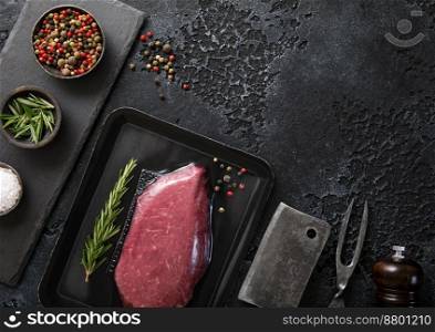 Raw beef fillet steak in vacuum tray with pepper on on black background with meat cleaver and fork. Top view.
