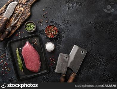 Raw beef fillet steak in vacuum tray with rosemary on on black background with meat cleavers and barbeque fork and knife.Top view.