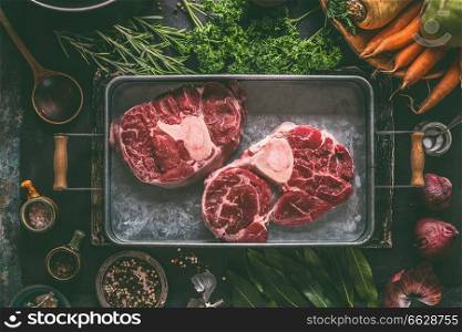 Raw beef meat shin with bone for broth, soup or stew in metal tray with ingredients for tasty cooking: root vegetables, herbs and spices on dark rustic kitchen table background, top view.