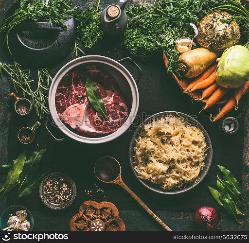 Raw beef meat shin with bone in cooking pot with spices and bowl with pickled cabbage on dark kitchen tables background with low carb ingredients, top view. Stew or meat soup preparation.