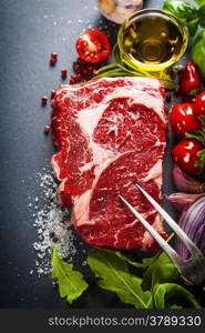 Raw beef steak with meat fork and ingredients on a dark slate background