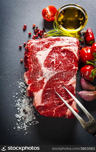 Raw beef steak with meat fork and ingredients on a dark slate background