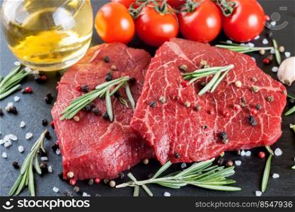 Raw beef steak with spices and ingredients for cooking on black slate background. Beef steak, rosemary, tomato, olive oil, garlic and spices. Food background.. Raw steak with cooking ingridients on black slate background