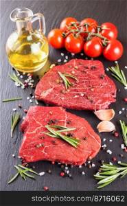 Raw beef steak with spices and ingredients for cooking on black slate background. Beef steak, rosemary, tomato, olive oil, garlic and spices. Vertical food background.. Raw steak with cooking ingridients on black slate background